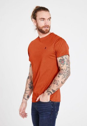 Roestkleurig katoenen t-shirt met korte mouwen en een ronde hals. Bevat een klein geborduurd logo op de borst. Geverfd met donkerblauwe spijkerbroek.