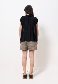 Schwarzes, gefaltetes Oberteil mit kurzen Ärmeln und kragen, kombiniert mit beigen und schwarzen karierter Shorts. Braune Schuhe mit Schnürsenkeln.