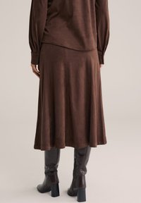 Femeie purtând un top maro cu mâneci lungi și o fustă midi asortată, cu cizme maro închis cu toc, până la genunchi, stând pe un fundal neutru.