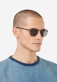 Mustad kaheksanurkse kuju ja tumedate läätsedega päikeseprillid, õhukeste metalle raamidega. Kandja on lühikese juuksega modell, kes kannab sinist denimit särki.