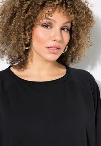 Top in tessuto nero e morbido con scollo ampio e maniche a raglan. Il modello presenta capelli ricci e orecchini a cerchio. Sfondo grigio chiaro.