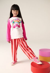 Valge pika varrukaga särk roosade varrukatega ja kirsside graafikuga, mida kannavad punaste triipudega heleroosad püksid ja roosad tossud.