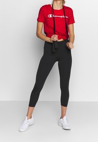 Rød kortærmet skjorte med hvidt logo, parret med sorte ankle-længde leggings og hvide sportssko, med et sjippetov i hånden.