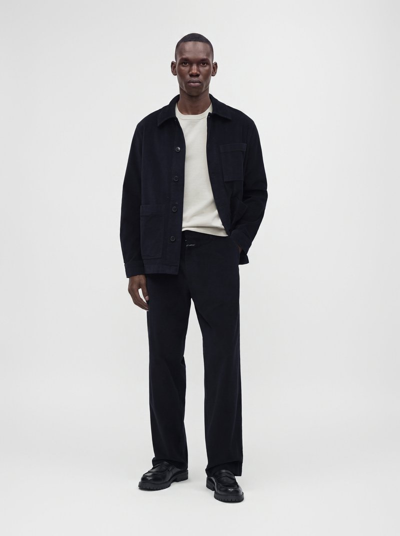 Homme debout devant un fond uni, portant une veste noire, une chemise de couleur claire, un pantalon noir et des chaussures noires, avec une main dans la poche.