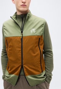 Ελαφρύ γιλέκο σε ελαιί πράσινο και καφέ με φερμουάρ μπροστά, υφή από δίχτυ και κεντημένο λογότυπο. Διαθέτει ψηλό γιακά και τσέπες χωρίς ραφές.