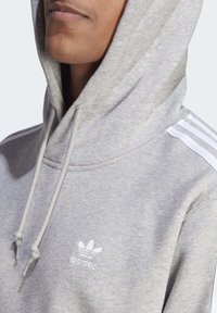 Grå huvtröja med vita dragsnören, har tre vita ränder på axlarna och en liten vit Adidas-logotyp på bröstet.