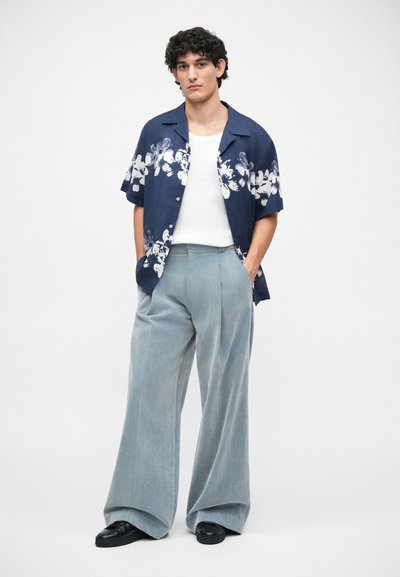 Chemise à manches courtes bleu marine avec un motif floral blanc, associée à un pantalon large bleu clair et des chaussures noires.