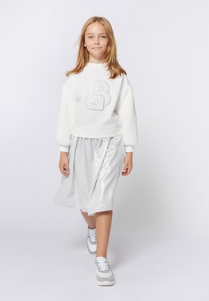 Sweatshirt blanc avec un logo noir structuré et des manches en peluche, associé à une jupe plissée argentée brillante et des baskets grises.