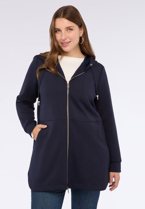 Donna che indossa una giacca a zip con cappuccio blu navy con tasche sopra una maglietta bianca e jeans blu, sorridendo con una mano in tasca.