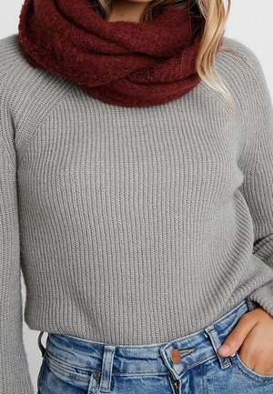 Femme portant un pull gris côtelé, un jean bleu et une épaisse écharpe infinie tricotée rouge foncé autour du cou.
