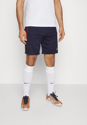 Homme portant des chaussettes blanches, des crampons noirs Nike, un short de football Paris Saint-Germain bleu marine et un t-shirt de sport blanc, debout devant un fond uni.