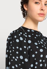 Blusa preta com bolinhas azul-claro, com mangas bufantes. Modelo usa grandes brincos de argola prateados. Tecido suave e leve.