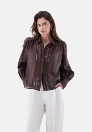 Frau mit langem braunem Haar, die goldene Creolenohringe trägt, eine braune Bluse mit Knöpfen und Spitzenverzierungen sowie weiße Hosen, Hände in den Taschen.