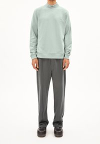 Vaaleanvihreä collegepaita korkealla kauluksella, yhdistettynä harmaisiin laskoshousuihin ja mustiin puukenkätyyppisiin kenkiin, joissa on sileät tekstuurit ja yksinkertainen muotoilu.