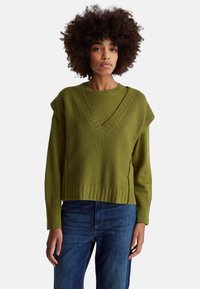 Olivově zelený pletený svetr se širokým výstřihem do V, žebrovanou strukturou a spadlými rameny, doplněný modrými džíny z denimu.