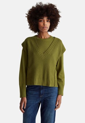 Olivgrüner Strickpullover mit weitem V-Ausschnitt, gerippter Textur und überschnittenen Schultern, kombiniert mit blauen Denim-Jeans.