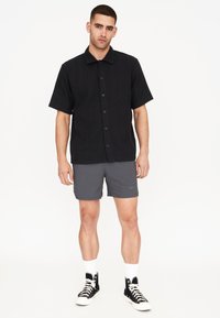 Maglietta nera a maniche corte con finitura texturizzata, chiusura a bottoni, abbinata a pantaloni corti grigi e sneaker alte nere con lacci bianchi.