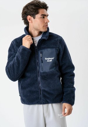 Homme portant une veste polaire bleu marine avec le texte « "Toujours Actif" », regardant sur le côté et tenant le col, fond clair.