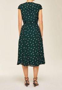 Robe verte avec un corsage ajusté, une jupe évasée et un motif de feuilles blanches. Manches courtes et taille définie. Portée avec des talons noirs à lanières.