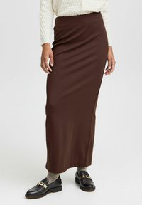 Gonna maxi aderente marrone realizzata in tessuto liscio, con una silhouette dritta. Abbinata a scarpe nere e calze texturizzate.