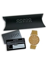 Montre OOZOO avec un cadran doré et des accents dorés, un bracelet en silicone beige, et une boîte noire avec une carte de garantie en dessous.