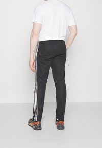 Černé sportovní kalhoty se třemi bílými pruhy po stranách, elastickým pasem a slim fit, doplněné černými botami s oranžovými akcenty.