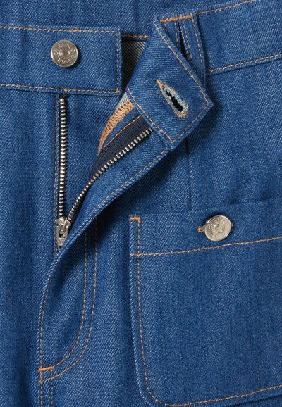 Pantalons en denim avec une fermeture éclair en métal, des coutures en cuivre et une poche avant avec fermeture à bouton en métal. Le tissu est d'un bleu moyen.