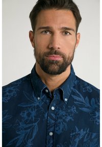 Camicia blu navy con bottoni, caratterizzata da un motivo floreale di un blu più chiaro, con colletto e taschino sul petto. La trama del tessuto appare liscia.