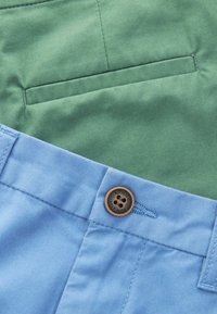 Tissu en coton bleu clair et vert avec un bouton portant le texte "AUTHENTIC NEXT DUDE". Texture lisse, couture précise et détail de poche.