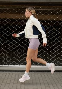 Hvit og marineblå zip-up jakke, tettsittende lavendel shorts og rosa sneakers med kraftige såler. Modell i bevegelse mot en bakgrunn med sekskantet mønster.