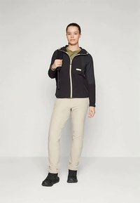 Chaqueta negra con cremallera y capucha, cremallera de contraste y acentos beiges. Combinada con pantalones beiges y calzado negro sobre un fondo neutro.