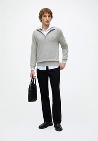 Jeune homme debout portant un pull gris clair à fermeture éclair quart sur une chemise blanche, pantalon noir et chaussures noires, tenant une mallette noire.