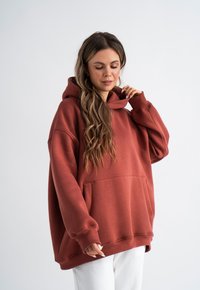 Oversized Kapuzenpullover in Rostfarbe, aus weichem Material mit einer Fronttasche und gerippten Bündchen. Getragen über weißen Hosen.