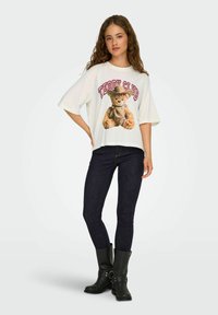 Biała, oversize'owa koszulka z grafiką misia teddy i napisem "TEDDY CLUB", w połączeniu z ciemnymi dżinsami i czarnymi kozakami do kolan.
