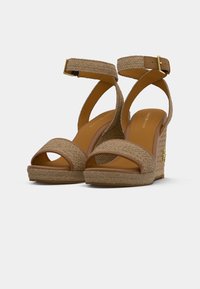 Sandales compensées tissées beiges avec des brides de cheville et des fermetures à boucle dorée, dotées d'une plateforme texturée et d'un design à bout ouvert.