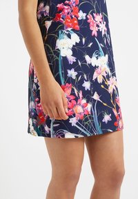 Květinové šaty s námořnickým pozadím, zdobené barevnými květinovými vzory v růžové, červené a bílé. Hladká textura s přiléhavým střihem.