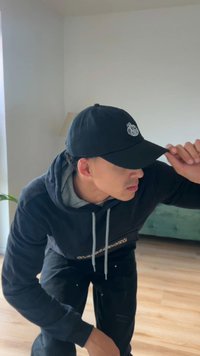 Fekete baseballsapka elöl logóval, szürke kapucnis pulóverrel és fekete nadrággal viselve. Világos fa padló és zöld kanapé látható a háttérben.