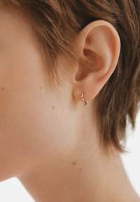 Pendiente de aro dorado con un pequeño acento de cristal, llevado en una oreja con cabello corto, resaltando un diseño simple pero elegante. Se aprecia el tono de piel.