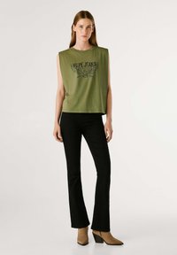 Maglietta senza maniche verde oliva con logo nero stampato; abbinata a pantaloni neri a zampa d'elefante e stivaletti beige, evidenziando un outfit casual.