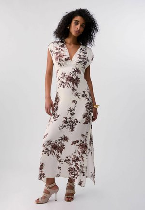 Donna con capelli ricci che indossa un lungo vestito bianco a fiori e sandali beige con tacchi alti, in piedi davanti a uno sfondo semplice.