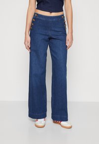 Pantalons en denim à jambes larges bleu foncé, avec une taille haute et six boutons décoratifs sur le côté gauche. Texture lisse, design décontracté.