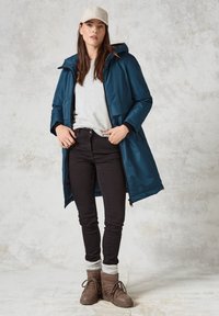 Teal vattentät jacka med huva, grå tröja, svarta jeans och bruna snörkängor med stickade ärmar, mot en ljus texturerad bakgrund.
