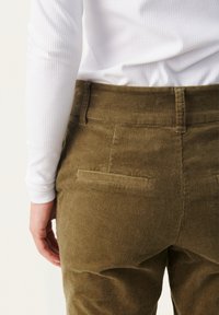 Pantaloni in corduroy verde oliva con tasche posteriori, abbinati a un top bianco a maniche lunghe con righe. Incentrati sulla texture e sul contrasto di colore.