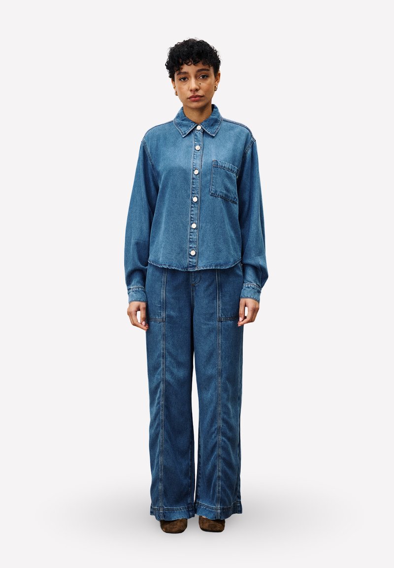 Vrouw staat, gekleed in een blauwe denim blouse met knopen en zak, en bijpassende denim broek met wijde pijpen, bruine schoenen zichtbaar.
