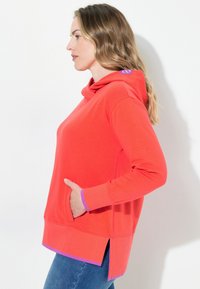 Sudadera roja con un bolsillo frontal, acentos morados en los puños y el dobladillo, y aberturas laterales. Fabricada con una tela suave y transpirable.