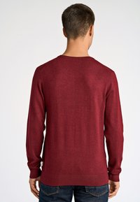 Bordowy sweter z dzianiny, z długimi rękawami i okrągłym dekoltem; ma elastyczne mankiety i dół, miękką teksturę oraz subtelny wzór.