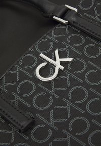 Černá kabelka s texturovaným vzorem a světle modrým nápisem, stříbrným logem na kovovém doplňku a hladkým, strukturovaným designem.