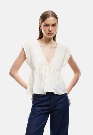 Ung kvinde med glat brunt hår har en ærmeløs hvid V-hals peplum top og mørkeblå jeans på, poserer med hænderne på hoften.