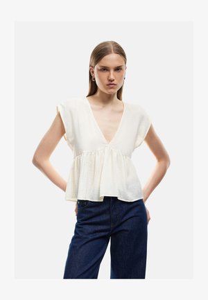 Giovane donna con capelli castani lisci indossa un top bianco senza maniche con scollo a V e peplum e jeans blu scuro, posando con le mani sui fianchi.