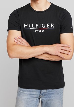 Mężczyzna z założonymi na piersi rękami, ubrany w czarny t-shirt z napisem "Hilfiger New York" oraz jasnoniebieskie dżinsy na jednolitym tle.